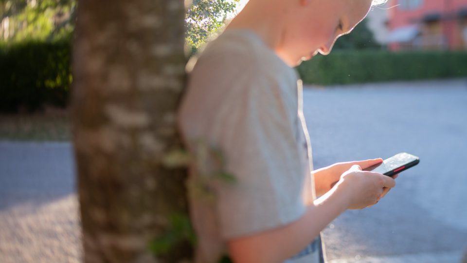 En pojke står lutad mot ett träd med en mobil i handen