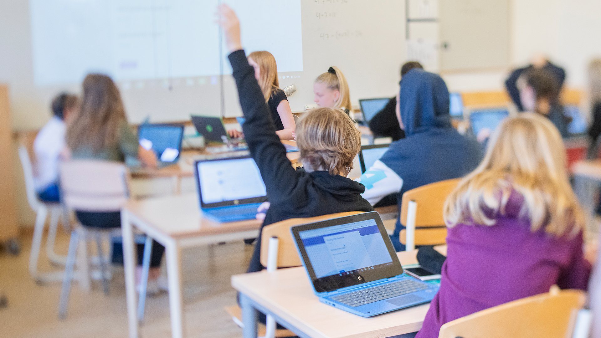 Klassrum där eleverna har laptops, en pojke räcker upp handen.