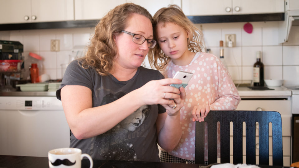 Kvinna visar ett barn något på mobiltelefon vid köksbord.