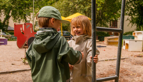 Två förskolebarn står i en lekpark vid en klätterställning.